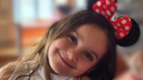 A close-up picture of Aria Thorpe. She is leaning her head to the side and she is smiling. On top of her head are Minnie Mouse ears.