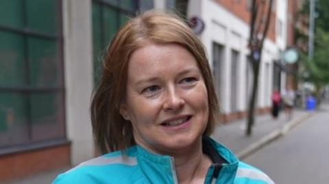 Anne has short ginger hair and is smiling at the camera as she looks to her left. She has a blue coat on, and behind her is brown and white buildings.