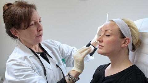 A woman is standing on the left wearing a white lab coat and has brown hair in a ponytail. She is holding a needle which she is injecting into a lady's face who is sat on the right, and who has blonde hair in a bun and sitting on a chair.