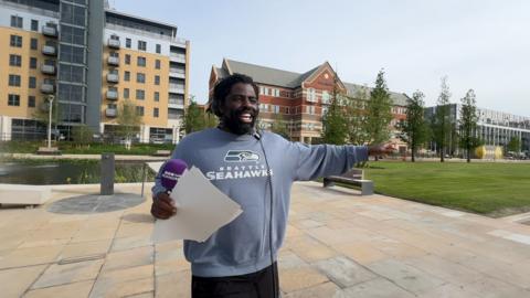 Person wearing a grey “Seattle Seahawks” sweatshirt and over‑ear headphones stands outdoors holding papers, with one arm outstretched. In the background, a modern public space features a tall white sculptural column, benches, greenery, and brick buildings under a blue sky.