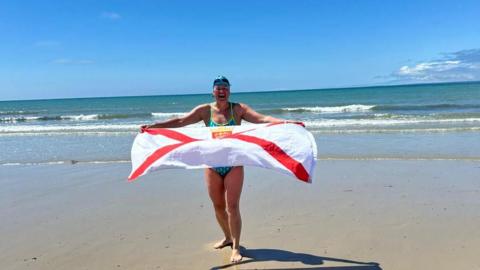 A woman holding a Jersey flag on the beach. Blue skies.