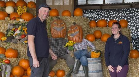 Man in blue t-shirt and blue cap and dark trousers. He is smiling. He is stood with a woman with sunglasses on her head, blue fleece, grey top and maroon trousers. She is smiling. There are lots of pumpkins in the background.