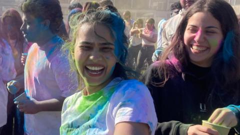 A woman is standing in the middle of the image, and has blue and green powder on her hair, face and white top. She is laughing at the camera. There are other people standing around her who are also covered in multiple coloured powders.