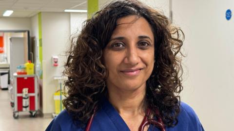 Dr Sarb Clare stands in the new Midland Metropolitan University Hospital. She has black, curly hair to her shoulders and half smiles at the camera. The red tubing of a stethoscope can be seen hanging around her neck along with the blue top of the scrubs she is wearing.