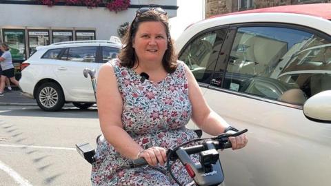 Anna Taylor on mobility scooter next to cars parked in Church Square in St Peter Port