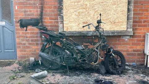 A heavily burnt out motorcycle leaning against a house