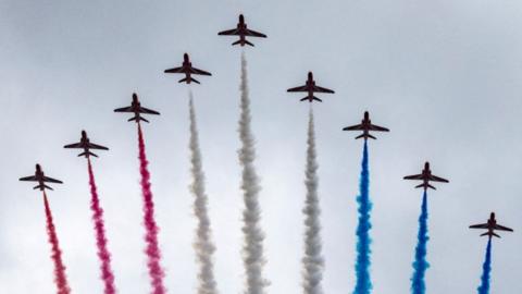Nine Red Arrows aircraft fly against a grey sky, trailing smoke coloured red, white and blue behind them.