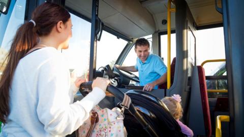 A bus driver in a blue polo shirt smiles at a child as her mother, in a white top, pushes her buggy on to a bus.