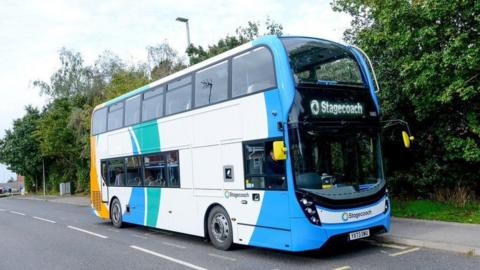 A Stagecoach East Midlands bus parked at a bus stop. The vehicle is a double decker painted in a white, blue, green and orange livery. It is on a grey tarmac road which is surrounded by leafy green trees.