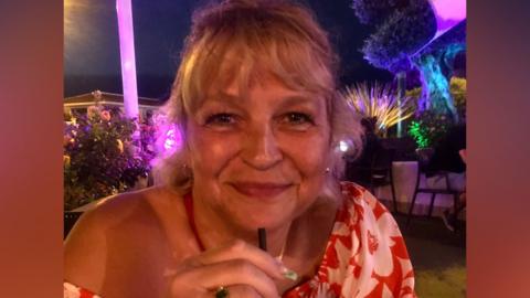 A person wearing a red‑and‑white patterned off‑the‑shoulder top is seated outdoors at night, holding a drink with a straw.