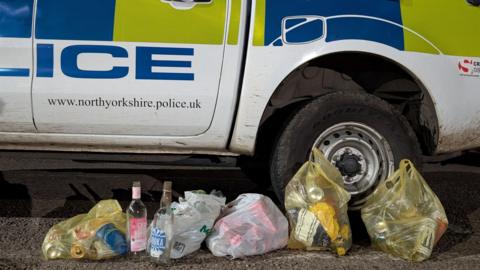 Five carrier bags of rubbish are on the ground by a police car, three yellow bags and two white ones. They are full of cans and glass bottles, with two glass bottles sitting in front of one of the bags.