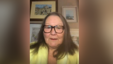 A woman takes a selfie wearing a yellow top with straight brown hair and large rimmed brown glasses. Behind her are framed photos of landscapes with ancient buildings.