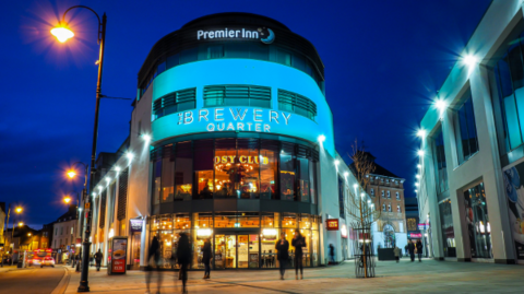 The Brewery Quarter in Cheltenham lit up at night. The main building shown in the picture is rounded at the corner facing the camera, and shows a cafe on the bottom floor, with a Cosy Club and a Premier Inn above it. People are walking by.