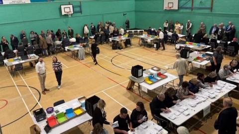 people are standing around a sports hall with people sitting at table and chairs counting votes.