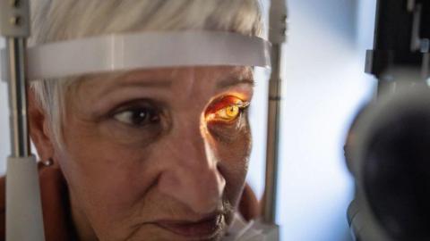 A woman has her forehead rested on a brace as a torch is shone into her left eye. 