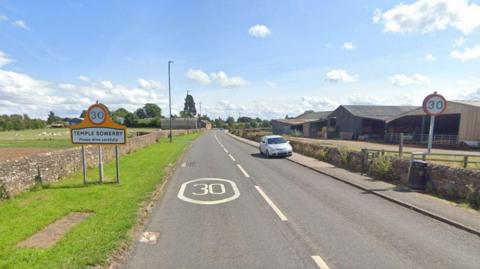 The B6412 road leading into Temple Sowerby. A sign on the right states the name of village as well as a 30 mph speed limit. A farm building stands on the right.