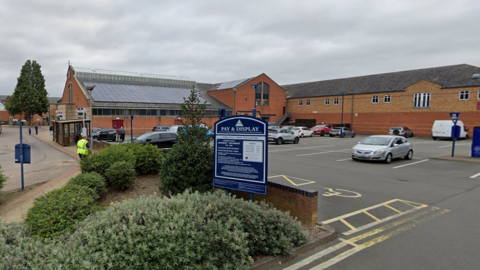 A council car park in Market Harborough next to the town's market hall. A number of cars are parked in spaces, and there is a shrubbery bed just outside the car park, and a large Pay & Display sign can be seen.