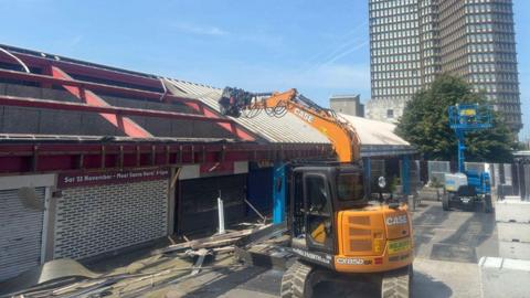 A yellow demolition vehicle removes corrugated roof panels from a single storey structure