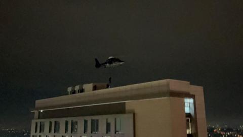 A helicopter is hovering above the roof of a cream-coloured multi-storey building, with a rope and someone dressed in black descending on to the roof. Some people in black can also be seen on the roof. City lights can be seen in the distance.