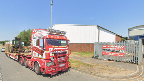 A red Scorpion Engineering truck outside a gate with a sign which says 'Scorpion Trailer Service Centre' - there are two buildings with corrugated wall panels.
