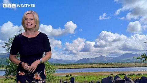 Sarah Keith-Lucas stands in front of a photograph of cloudy blue sky, over mountains and a lake