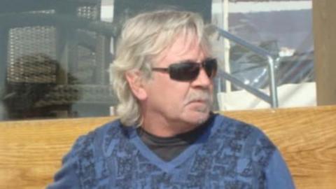 Michael Shirley has collar-length grey hair and is wearing black sunglasses and a blue V-neck jumper over a black T-shirt. He is looking to the side while sitting on a wooden bench.