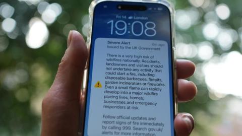 A hand holding an iPhone which includes a notification of an emergency alert from the UK Government on wildfires.