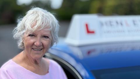 Olwen has white short hair. She smiles for the camera in front of her learner car. She is wearing a pink sweater.
