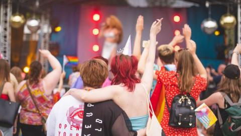 A crowd of people stood in front of a stage. Some people are holding rainbow flags. Others are holding their hands up in the air.