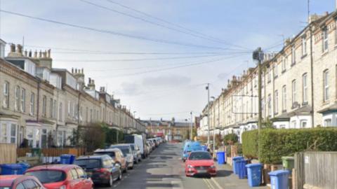 A street with terraced houses on both sides, cars parked on both sides and blue wheelie bins outside each property.