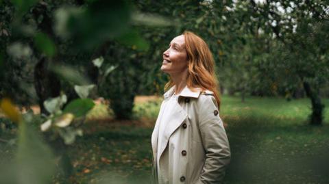 A woman is in a park surrounded by trees and looking happy. She has long hair and is wearing a beige coat and is looking up at the sun.