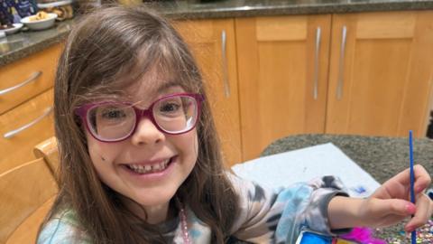 Saffie, wearing purple glasses, holding a paint brush in one hand. She has on purple glasses and has long dark hair. She is sitting by a table in a kitchen, with cabinets all around her.