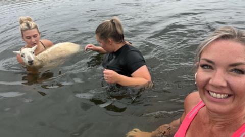 Aimee Turpie, who is wearing a pink running top smiles broadly as she takes a selfie to show Britney Lawrence and Abi Cox rescuing a sheep in Siblyback Lake in Cornwall. Britney is carrying the sheep in the water. Abi is stood next to her and is wearing a black top. All three are either waist or chest deep in the water.