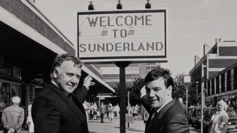 A black and white image of two men in suits pointing to a sign that reads: "Welcome to Sunderland". They are standing on a high-street bordered by shops in concrete and brick buildings. The men are smiling at the camera.