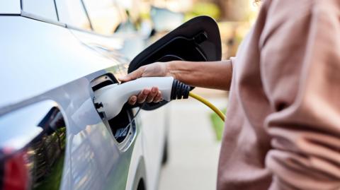 A mid section shot showing the body work of a silver car and a person wearing a salmon pink jumper fitting the nozzle of an EV charger into the vehicle's charging port.