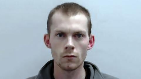 Close-up portrait of Connor Manners. He is facing the camera against a plain light background, with very short hair and wearing a grey top.