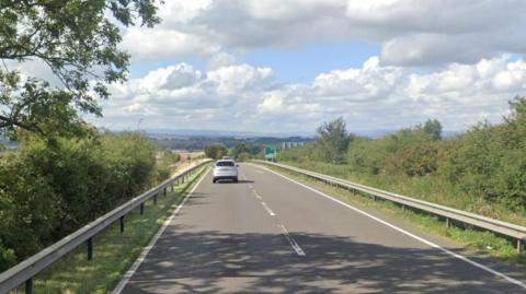 A dual carriageway road with trees on either side and a view of hills in the distance. A silver car on the road. Blue sky with clouds.