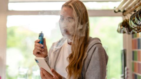 A young fair-haired woman wearing a grey hooded sweatshirt and white T shirt is standing in a kitchen with vape and mobile phone in her hands