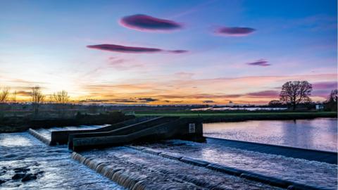 Purple lenticular clouds in a blue sky with an orange band over the horizon, above a weir