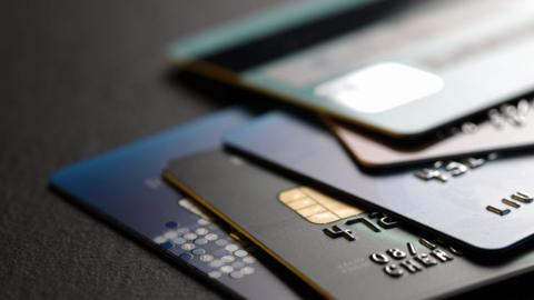 A close-up view of several payment cards in shades of blue, black and grey is shown fanned out on a dark surface. The cards display embossed numbers and metallic chip details.