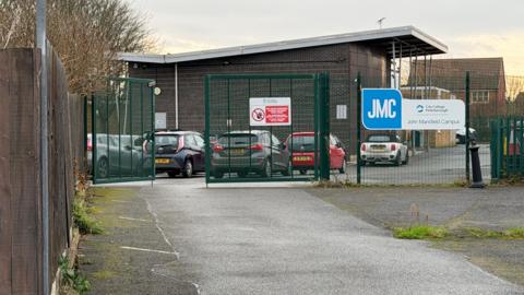 A general view of the John Mansfield Centre. There are cars in a car park in the foreground, behind a wire fence, and the centre building is in the background. It is a dark brick structure with a white roof.