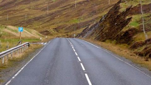 A road in Shetland with a parking sign to the left. There is banked grassland at both sides and another road leading off to the left after the parking sign