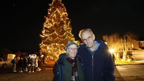 A elderly couple pose for a photo stood next to each other with a large Christmas tree behind them illuminated in warm lighting. A crowd of people are gathered behind them looking at the tree. 