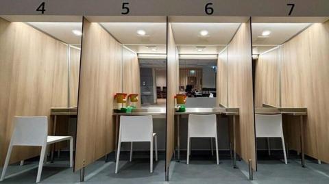 Numbered booths with a table and chair each are in a row. There is a waste bin in the foreground.
