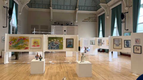 An art exhibition displayed in a large hall, with paintings and sculptures arranged on freestanding white panels and plinths.