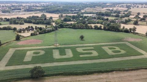 Aerial view of countryside with trees and hedges and green and yellow fields. The letters P U S B are cut into the nearest field.