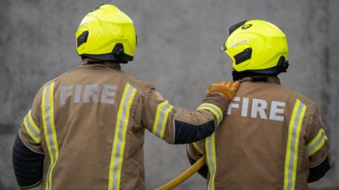 Two firefighters are wearing protective brown clothing with silver and yellow reflective strips. They have yellow helmets on their heads and one is wearing a brown glove with their hand on the other's shoulder. The other firefighter has a yellow hose