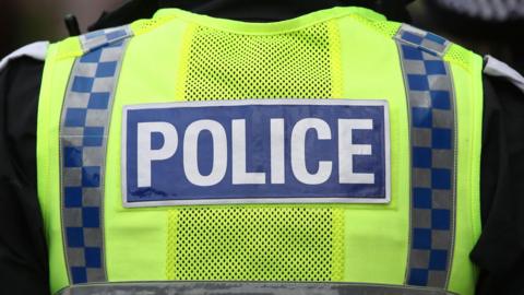 A police officer's upper back. Their vest is neon yellow with a blue label rectangular label with "police" written on it in white capital letters.