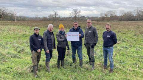 Ben Morgan-Brown, Councillor Geoff Jung, Councillor Paula Fernley, Councillor Henry Massey, Paul Osborne and James Chubb pictured standing in a large grassy field. They are wearing warm clothing and wellington boots while holding a large sheet of paper that reads Clyst Meadows.