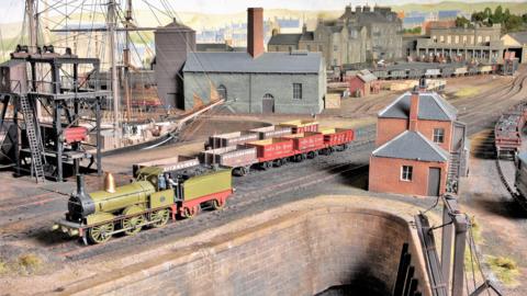 A model railway exhibition which is based on Burntisland in Scotland. It shows a busy railway site from the Victorian era with a green train in the foreground and a number of buildings behind.